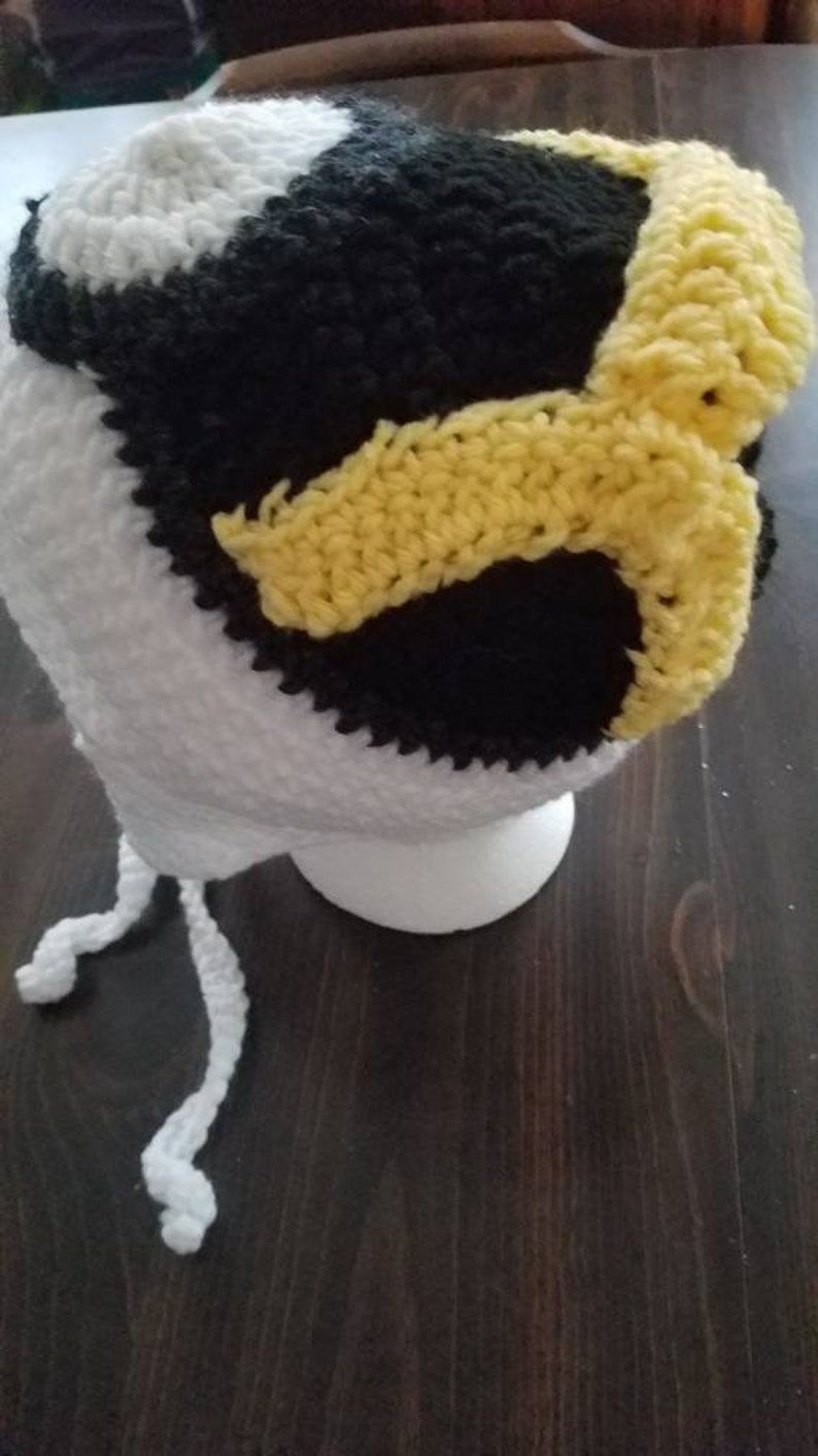 May include: White crocheted hat with black and yellow accents. The hat has a long white tie.