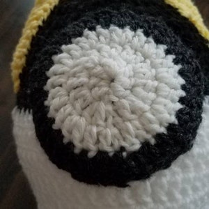 May include: A crocheted hat with a yellow, black, and white color scheme. The hat has a circular design with a white center, a black ring around it, and a yellow brim.