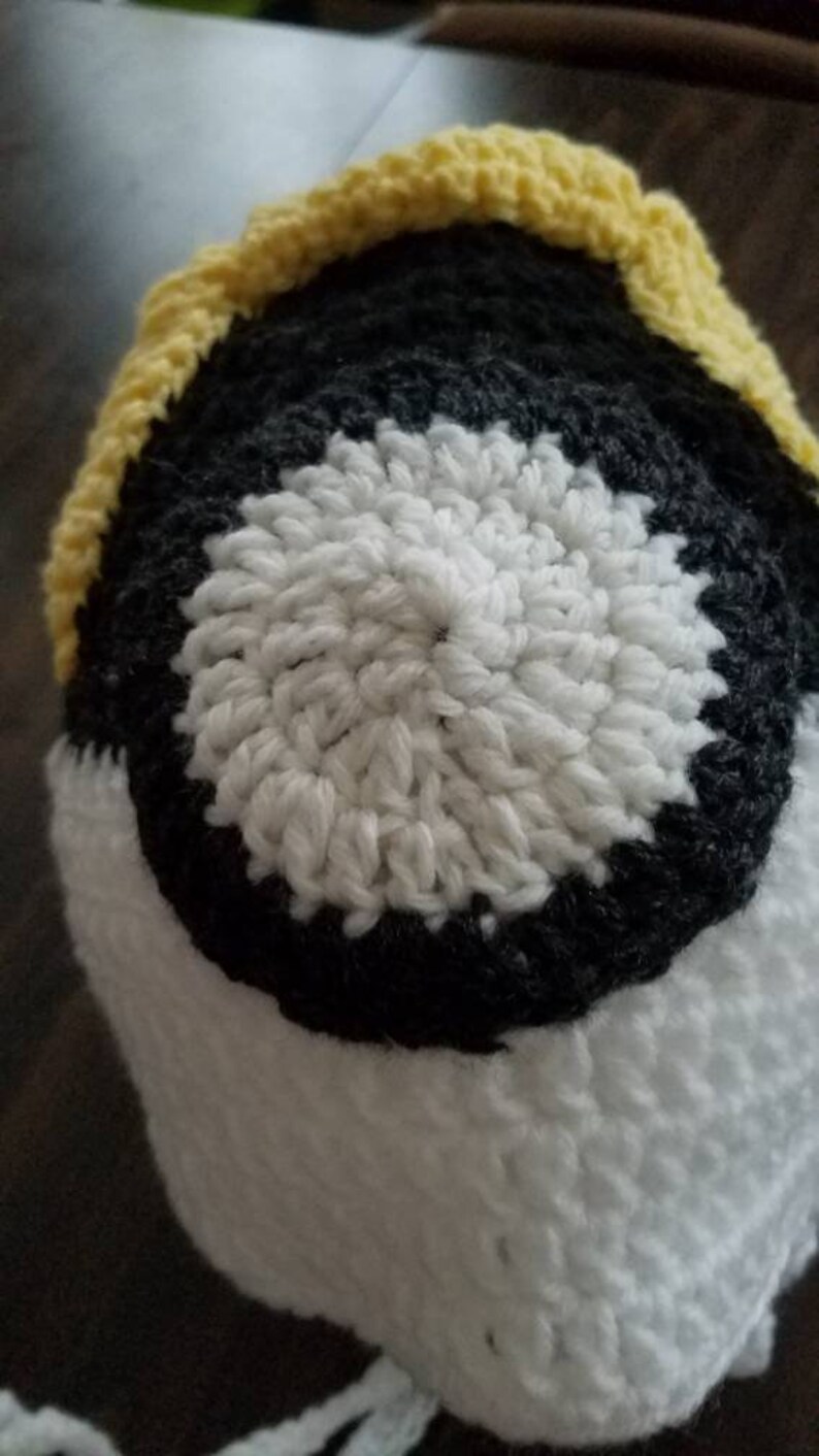 May include: A crocheted hat with a yellow, black, and white color scheme. The hat has a circular design with a white center, a black ring around it, and a yellow brim.