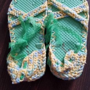 Op de afbeelding: Een paar groene slippers met gele, blauwe en groene gehaakte versieringen en groene kant.