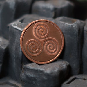 May include: A copper-colored coin with a Celtic knot design. The coin is sitting on a textured black surface.