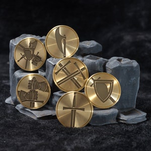 May include: Six gold-colored metal coins with different designs, including a sword, a shield, and a cross. The coins are arranged on a black surface.