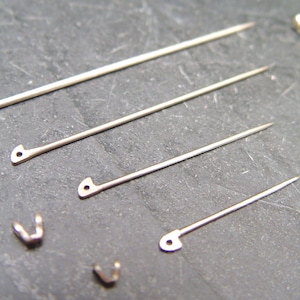 May include: Close-up of various silver-toned jewelry findings, including long pins with decorative heads, pins with eyelets, and small V-shaped clasps. The components are arranged on a dark, textured surface.