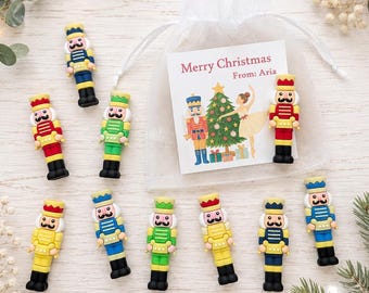 May include: A set of ten small nutcracker figurines in various colors including red, blue, green, and yellow, each with black boots and a mustache. A white organza drawstring bag features a card with the text Merry Christmas From: Aria.
