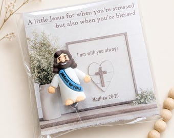 May include: A small figurine of Jesus with brown hair and a white robe featuring a blue sash that says Jesus loves you. It is packaged in a clear plastic sleeve with a card that reads A little Jesus for when you're stressed but also when you're blessed and I am with you always Matthew 28:20.
