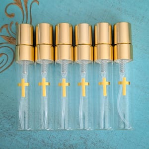 Puede incluir: Seis frascos pulverizadores de vidrio transparente dispuestos en fila, cada uno con un tap&oacute;n de color dorado y un peque&ntilde;o emblema de cruz dorada impreso en el frente. Los frascos tienen forma cil&iacute;ndrica con un mecanismo de pulverizaci&oacute;n visible.