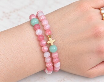 May include: Two beaded bracelets featuring round pink and light green stones. One bracelet includes a small gold-toned cross charm positioned between the beads. The bracelets are worn together on a wrist, showcasing the smooth, polished texture of the beads.