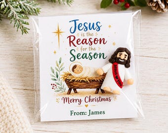May include: A small figurine of a bearded figure in a white robe with a red sash reading Jesus Loves You, attached to a card featuring a manger scene, the text Jesus is the Reason for the Season, Merry Christmas, and From: James in a clear plastic sleeve.