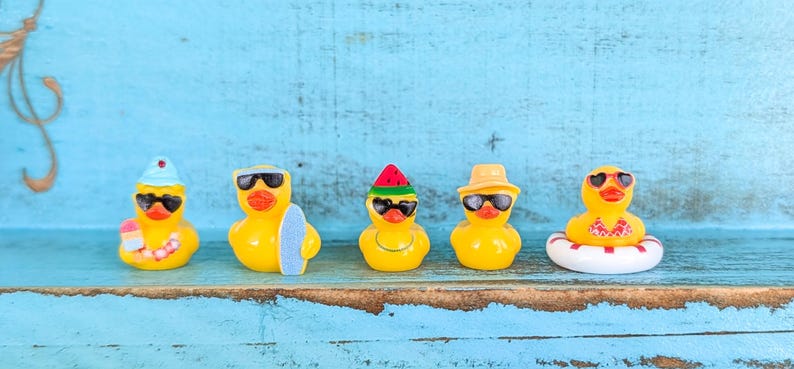 May include: Five yellow rubber ducks arranged in a row. The first wears a blue hat and sunglasses, the second has sunglasses and a surfboard, the third wears a watermelon hat and sunglasses, the fourth has a tan hat and sunglasses, and the fifth wears heart-shaped sunglasses and sits in a white and red life ring.
