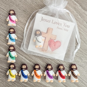 May include: A collection of small Jesus figurines with colorful sashes, alongside a sheer white bag containing a card that reads "Jesus Loves You." The card features a cross and a heart illustration. The figurines have brown hair and beards.