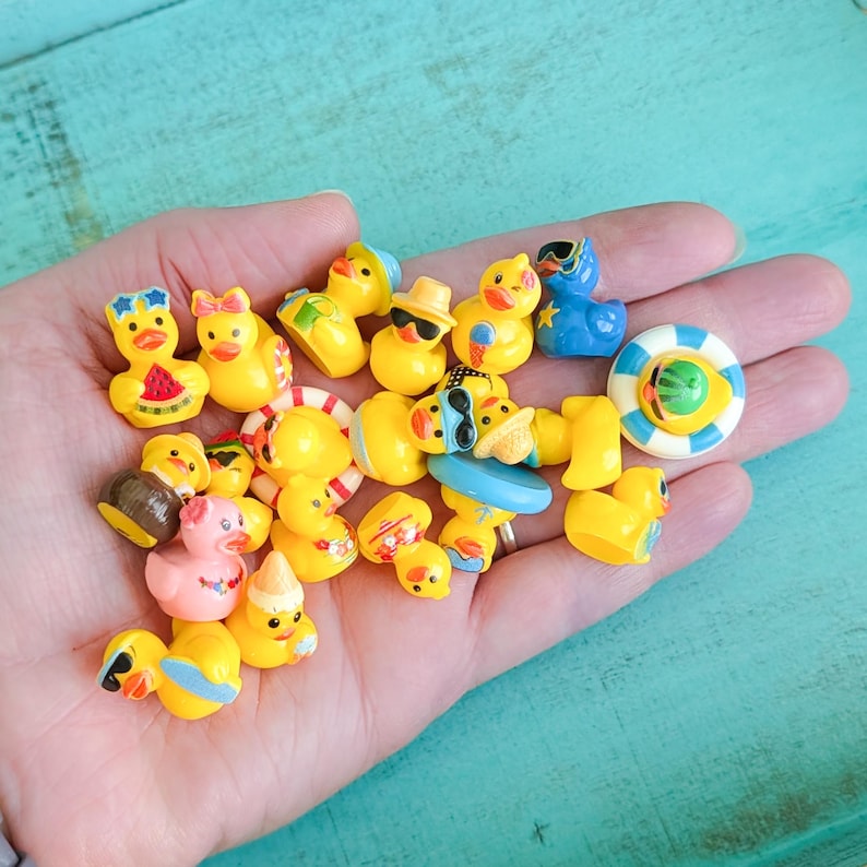 May include: A collection of miniature rubber ducks in various colors and designs, including some wearing sunglasses, hats, or holding small items like watermelon slices and ice cream cones, alongside a small blue and white striped life preserver.