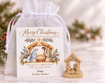 Puede incluir: Una bolsa de organza blanca transparente con cordón que contiene una tarjeta con una ilustración de la natividad y el texto Merry Christmas, A little reminder of the greatest gift of all, For unto us a child is born, unto us a son is given, Isaiah 9:6, From: James, y una pequeña figura de natividad beige.