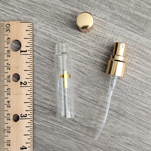 Puede incluir: Un frasco atomizador de vidrio transparente con un emblema de cruz dorada, una tapa dorada y un mecanismo de bomba rociadora dorado. Una regla de madera est&aacute; junto al frasco, mostrando que el recipiente de vidrio mide unos 6,4 cm de alto.