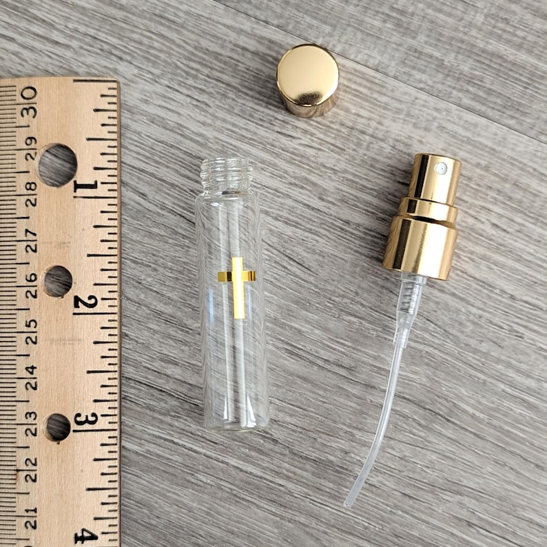 Puede incluir: Un frasco atomizador de vidrio transparente con un emblema de cruz dorada, una tapa dorada y un mecanismo de bomba rociadora dorado. Una regla de madera est&aacute; junto al frasco, mostrando que el recipiente de vidrio mide unos 6,4 cm de alto.