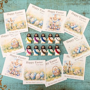 May include: An assortment of Easter-themed greeting cards and small Jesus figurines. The cards feature watercolor illustrations of crosses, Easter eggs, and lambs, with the text "Happy Easter" and "He has risen!". The figurines are white with colorful sashes.