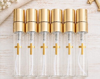 May include: Six clear glass spray bottles with gold-colored caps and a gold cross emblem printed on the front of each bottle. The bottles are arranged in a row, each featuring a clear plastic dip tube inside and a metallic gold spray nozzle mechanism.