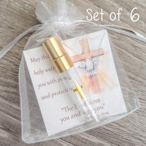 Puede incluir: Un frasco pulverizador de vidrio transparente con tapa dorada dentro de una bolsa de organza blanca. Una tarjeta interior muestra una cruz de madera, una paloma blanca y el texto: May this holy water bring you peace and protection.