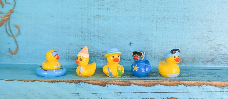 May include: Five small rubber duck figurines arranged in a row. The first is yellow with goggles in a blue inner tube. The second is yellow with a tan party hat. The third is yellow with a blue hat and bucket. The fourth is blue with goggles. The fifth is yellow with blue sunglasses.