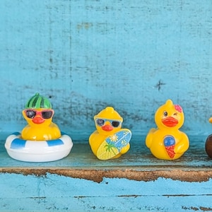 May include: Five small yellow rubber ducks arranged in a row. The first wears a floral lei, the second wears sunglasses and a watermelon hat in a blue and white inner tube, the third holds a surfboard, the fourth holds an ice cream cone, and the fifth wears a straw hat in a brown bowl.