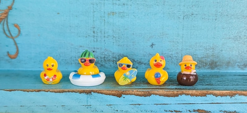 May include: Five small yellow rubber ducks arranged in a row. The first wears a floral lei, the second wears sunglasses and a watermelon hat in a blue and white inner tube, the third holds a surfboard, the fourth holds an ice cream cone, and the fifth wears a straw hat in a brown bowl.
