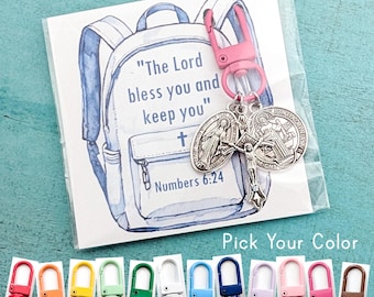 May include: A keychain featuring a pink lobster clasp and three silver-toned religious charms, including a crucifix and two medals, attached to a card with a watercolor backpack illustration and the text The Lord bless you and keep you.