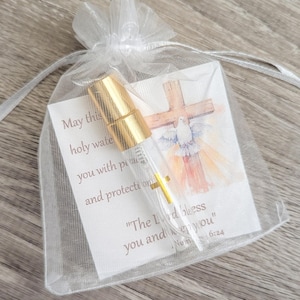 Puede incluir: Un frasco de vidrio transparente con tapa de spray dorada dentro de una bolsa de organza blanca con cord&oacute;n. El frasco descansa sobre una tarjeta con una cruz en acuarela, una paloma y el texto: May this holy water fill you with peace and protection. The Lord bless you and keep you. Num 6:24.
