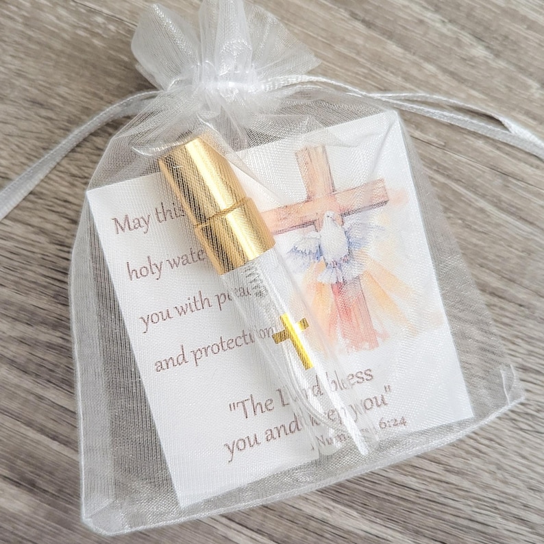 Puede incluir: Un frasco de vidrio transparente con tapa de spray dorada dentro de una bolsa de organza blanca con cord&oacute;n. El frasco descansa sobre una tarjeta con una cruz en acuarela, una paloma y el texto: May this holy water fill you with peace and protection. The Lord bless you and keep you. Num 6:24.