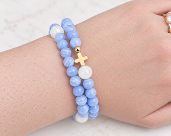 May include: Two stretch bracelets featuring light blue banded agate beads and white translucent beads. One bracelet includes a small gold-toned cross charm and a gold-toned spacer bead. The bracelets are worn together on a wrist.