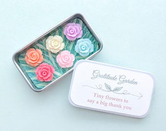May include: A rectangular metal tin containing six small, pastel-colored rose-shaped ornaments resting on a green leaf-patterned base. The lid features the text Gratitude Garden and Tiny flowers to say a big thank you with a leaf graphic.