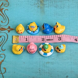May include: A collection of eight miniature rubber ducks in various colors and designs, including some wearing sunglasses or hats and others in floaties, arranged next to a pink measuring tape showing a length of approximately 3.5 inches.