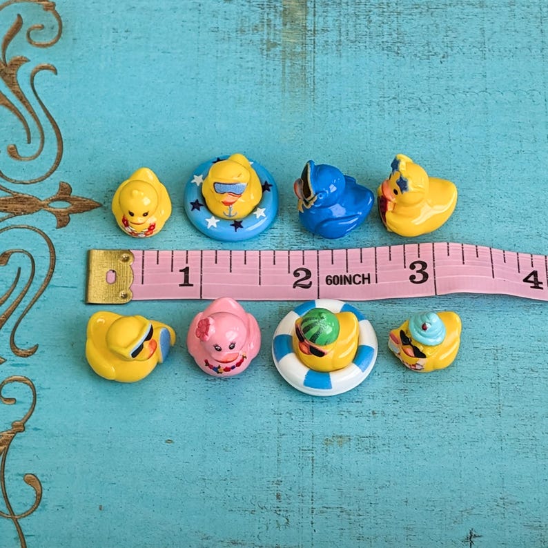 May include: A collection of eight miniature rubber ducks in various colors and designs, including some wearing sunglasses or hats and others in floaties, arranged next to a pink measuring tape showing a length of approximately 3.5 inches.