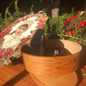 May include: A wooden oval-shaped box with a ring inside, set against a backdrop of greenery and a vintage portrait. The ring features a large, clear gemstone. The box is light brown with a lid and decorative rivets.