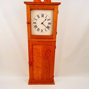May include: A tall, wooden grandfather clock with a warm, reddish-brown finish. The clock features a square face with Roman numerals and black hands. The clock has a cabinet-style base and a decorative top with a circular cutout.