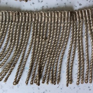 May include: A close-up of a silver fringe trim with a woven texture. The fringe is made of twisted strands of yarn and has a subtle sheen.