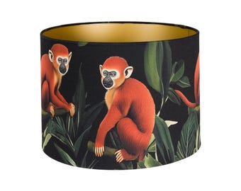 Könnte beinhalten: Zylindrischer Lampenschirm mit einem sich wiederholenden Muster aus orangefarbenen Affen und grünen Blättern auf schwarzem Hintergrund. Die Innenseite ist goldfarben. Das Design erzeugt ein tropisches Dschungelthema.