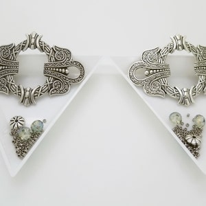 May include: Two ornate silver-colored metal clasps with intricate designs, each resting on a white triangular tray. Small beads and decorative elements in silver and pale green are also present, suggesting jewelry components or craft supplies.