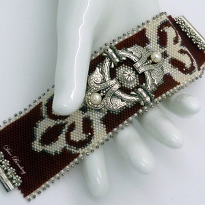 K&ouml;nnte beinhalten: Ein braunes und wei&szlig;es Perlenarmband mit einem silbernen Verschluss und zwei Perlenakzenten. Das Armband liegt auf einer wei&szlig;en Oberfl&auml;che.