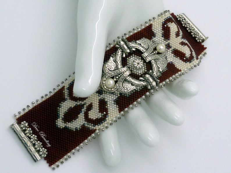 K&ouml;nnte beinhalten: Ein braunes und wei&szlig;es Perlenarmband mit einem silbernen Verschluss und zwei Perlenakzenten. Das Armband liegt auf einer wei&szlig;en Oberfl&auml;che.