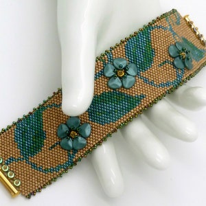 Könnte beinhalten: Ein Armband mit Perlen und einem floralen Muster. Das Armband ist aus braunen, grünen und blauen Perlen gefertigt. Das Armband hat einen goldenen Verschluss.