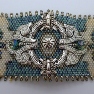 K&ouml;nnte beinhalten: Ein verzierter, dekorativer Verschluss in Silberton mit einem Perlenrand und zwei irisierenden blauen Edelsteinen. Der Verschluss ist zentriert auf einem Perlenarmband mit einem blauen, wei&szlig;en und beigefarbenen geometrischen Muster. Das Armband ist mit "Deco Beading" beschriftet.