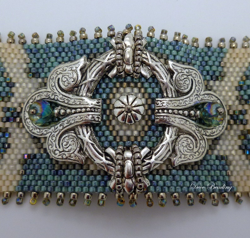 K&ouml;nnte beinhalten: Ein verzierter, dekorativer Verschluss in Silberton mit einem Perlenrand und zwei irisierenden blauen Edelsteinen. Der Verschluss ist zentriert auf einem Perlenarmband mit einem blauen, wei&szlig;en und beigefarbenen geometrischen Muster. Das Armband ist mit "Deco Beading" beschriftet.