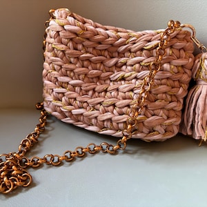 May include: A dusty pink crocheted handbag featuring a thick, chunky knit texture interwoven with metallic gold thread. The bag includes a long, polished copper-toned circular link chain strap and a matching fabric tassel attached to the side.