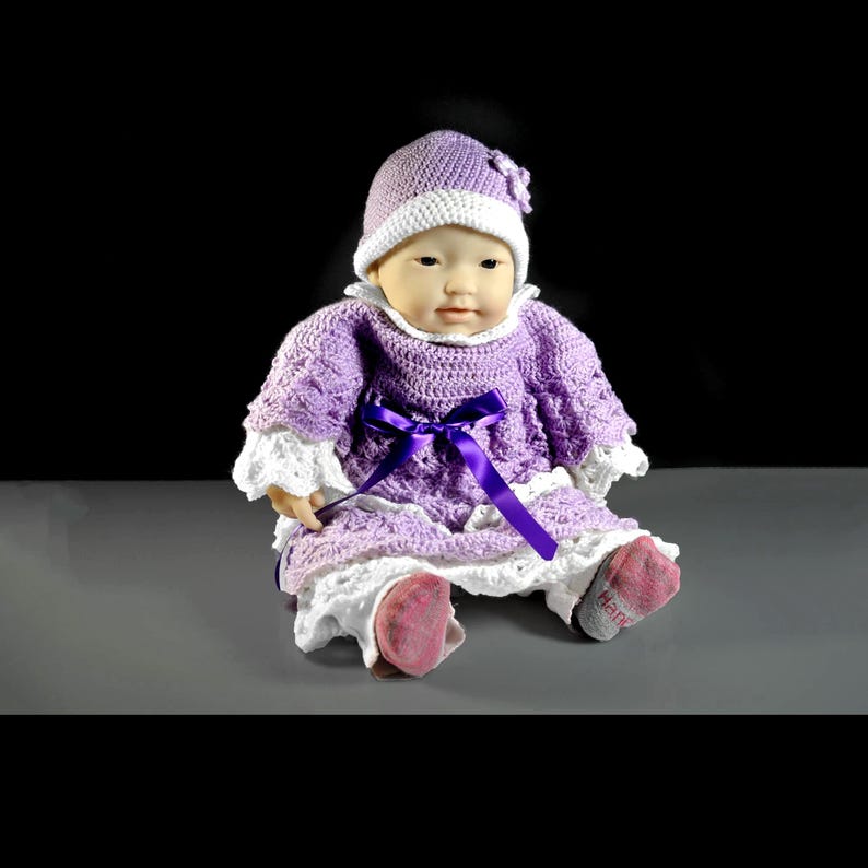 May include: A doll wearing a lavender crocheted dress with white lace trim at the sleeves and hem, a matching lavender crocheted hat with a white brim, and a dark purple ribbon tied at the waist. The doll has pink socks on its feet.