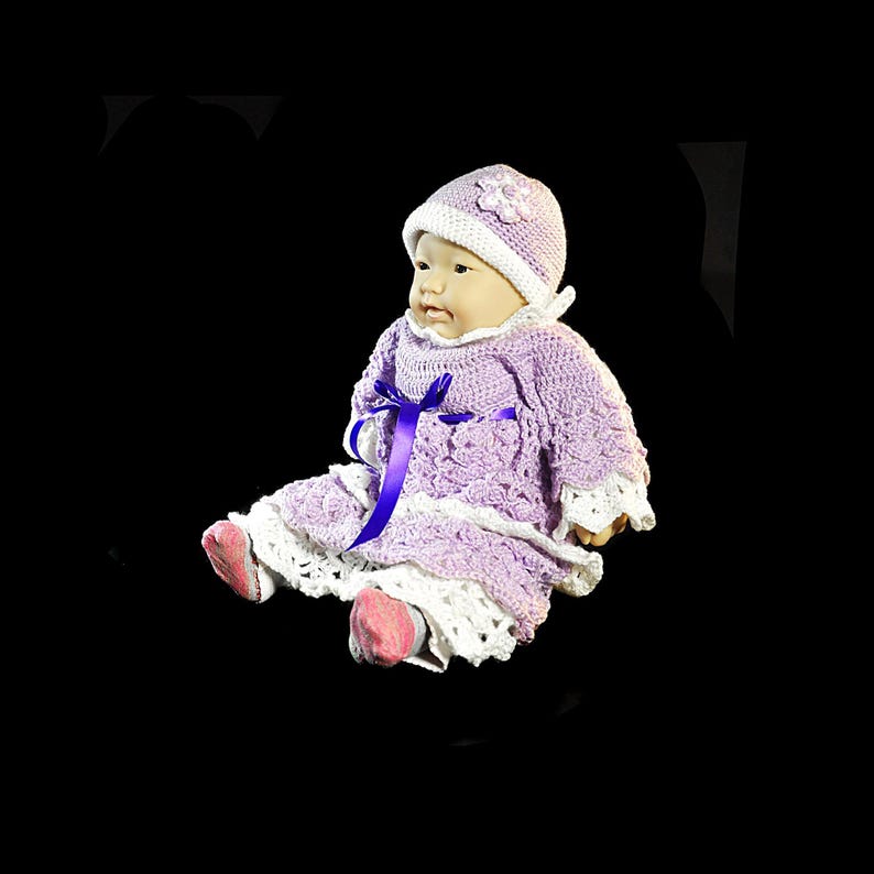 May include: A doll wearing a lavender and white crocheted dress with a matching lavender hat. The dress features a white crocheted hem, a purple ribbon tied at the waist, and a white collar. The hat is adorned with a small white flower embellishment.