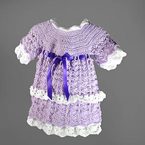 May include: A lavender crochet dress featuring a round neckline, short sleeves with white scalloped trim, and a tiered skirt with white crochet borders. A dark purple satin ribbon is tied in a bow at the waist, accentuating the textured pattern.