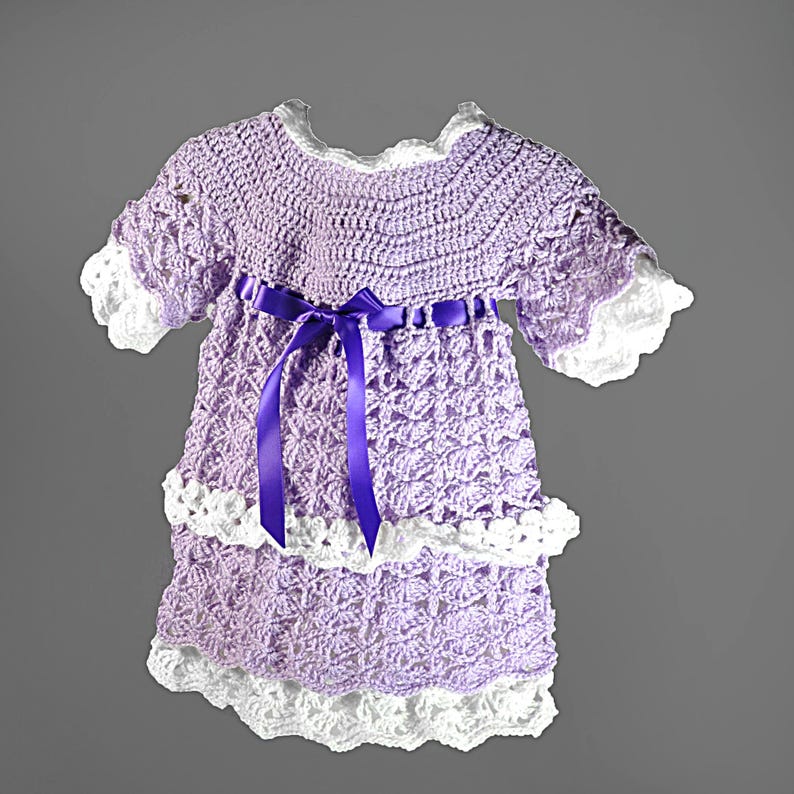 May include: A lavender crochet dress featuring a round neckline, short sleeves with white scalloped trim, and a tiered skirt with white crochet borders. A dark purple satin ribbon is tied in a bow at the waist, accentuating the textured pattern.