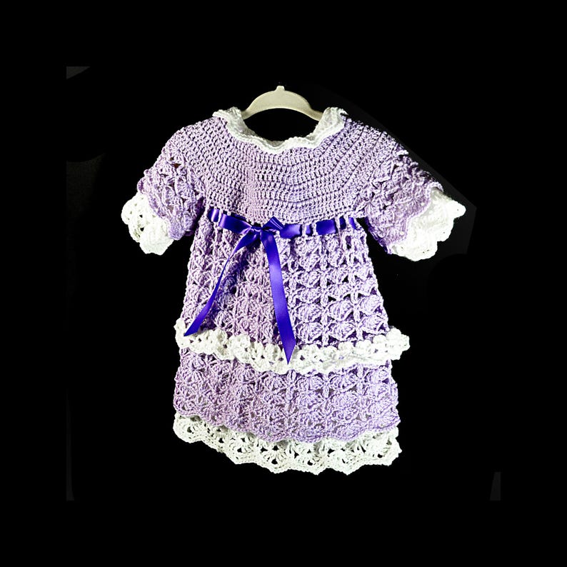 May include: A lavender crochet dress featuring a white scalloped collar and sleeve cuffs, a tiered skirt with white crochet trim, and a dark purple satin ribbon tied in a bow at the waist. The garment is displayed on a white plastic hanger.