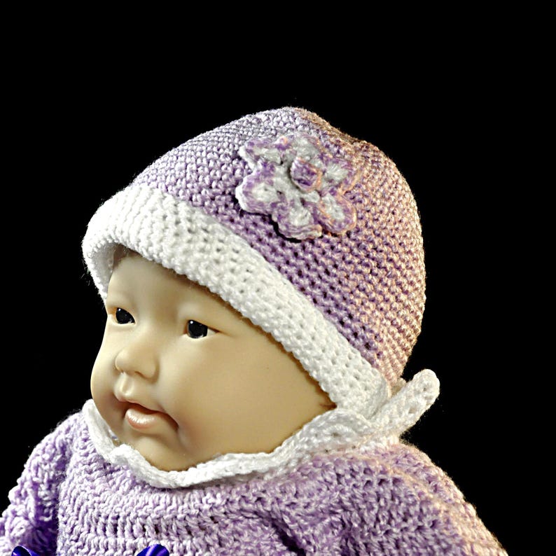 May include: A doll wearing a light purple crocheted bonnet with a white trim and a white crocheted flower accent on the side. The bonnet features white ties that are knotted under the chin. The doll is also wearing a matching light purple crocheted top.