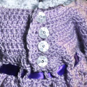 May include: A lavender crocheted garment featuring a white crocheted collar and four round, white, iridescent buttons with central clear crystals. A dark purple satin ribbon is woven through the crochet stitches near the bottom edge.