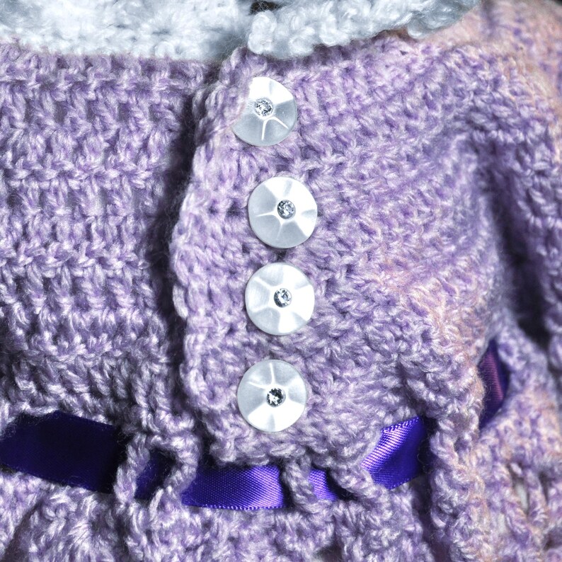 May include: A lavender crocheted garment featuring a white crocheted collar and four round, white, iridescent buttons with central clear crystals. A dark purple satin ribbon is woven through the crochet stitches near the bottom edge.